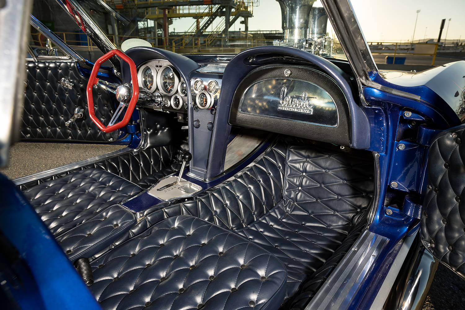 Wide view of the cockpit showing custom button-tufted dark blue vinyl upholstery, red steering wheel, and Corvette Sting Ray glovebox door.