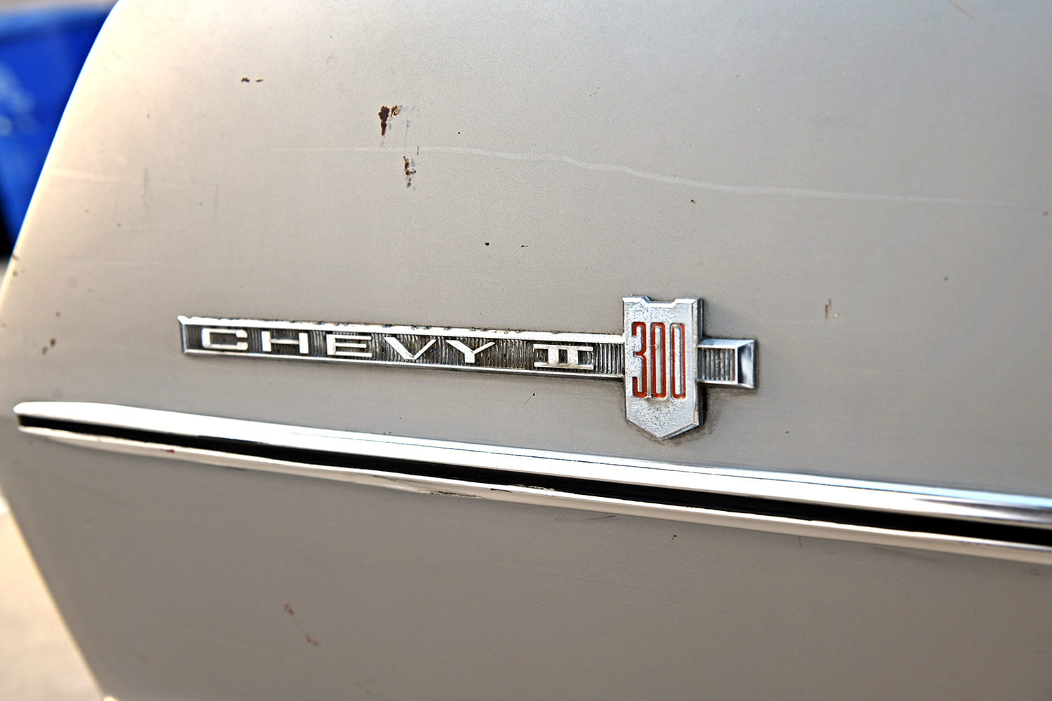 Macro view of the original die-cast chrome "Chevy II 300" emblem with red inlay mounted on the patina silver rear quarter panel.