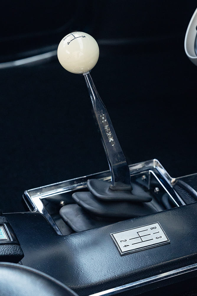 Console view focusing on the chrome Hurst four-speed manual shifter handle with a classic cream-white cue-ball shift knob and reverse lockout pattern.