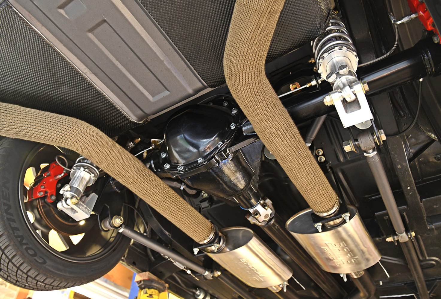 Undercarriage view of the rear axle showing coilover shocks, multi-link suspension arms, differential, and heat-wrapped dual exhaust pipes with stainless mufflers.