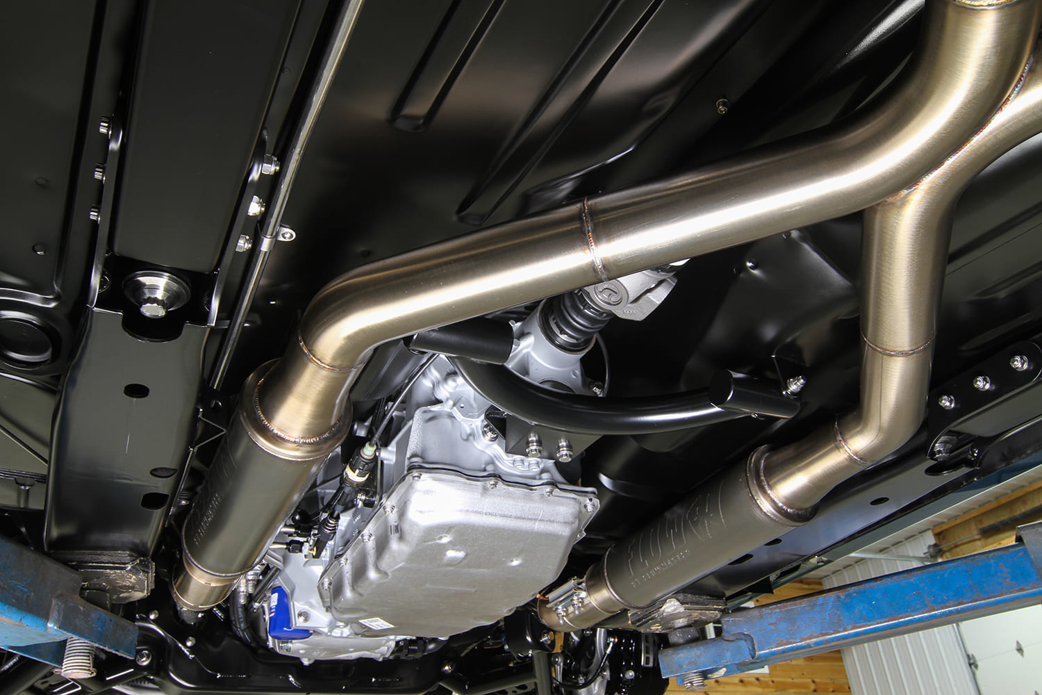 Underside view of the car on a lift displaying the transmission, driveshaft safety loop, and dual stainless steel exhaust system with mufflers.