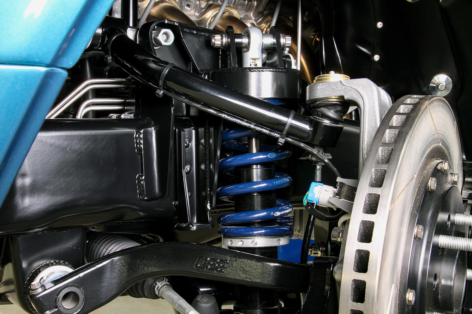 Front suspension detail showing blue coilover springs, tubular upper and lower control arms, and cross-drilled disc brake rotor.