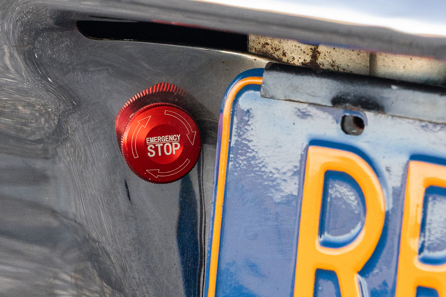 Close-up of a red anodized rotary master battery disconnect switch labeled "EMERGENCY STOP" mounted near the rear license plate bracket.