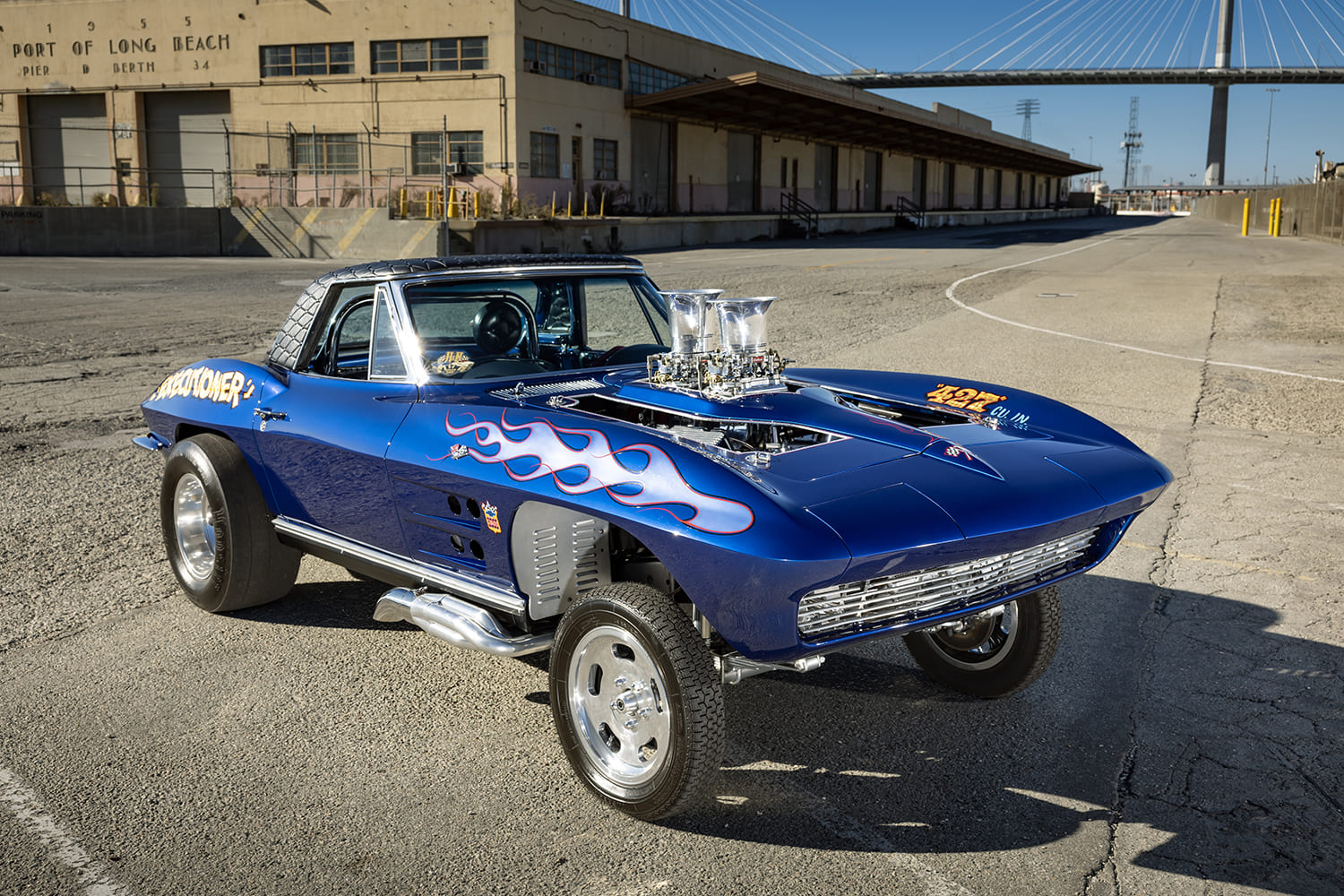 Elevated front three-quarter view of a blue 1964 Corvette gasser parked on open asphalt at the Port of Long Beach.