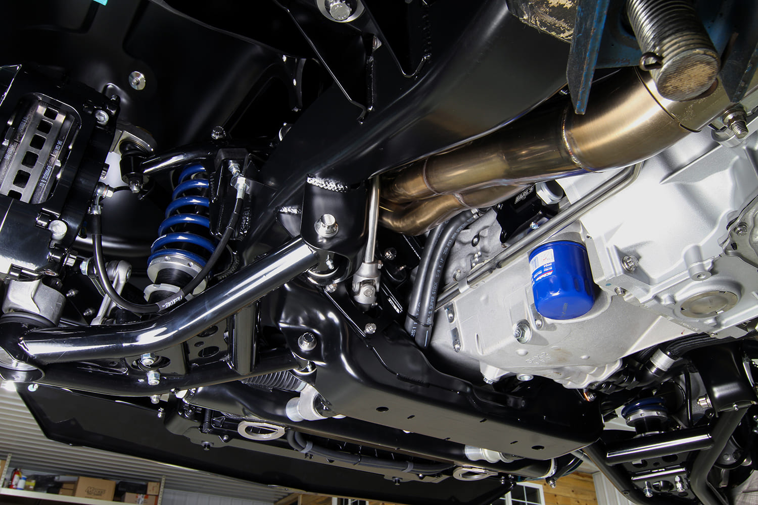 Lower suspension view showing the tubular lower control arm, blue coilover assembly, big brake caliper, and stainless exhaust header.