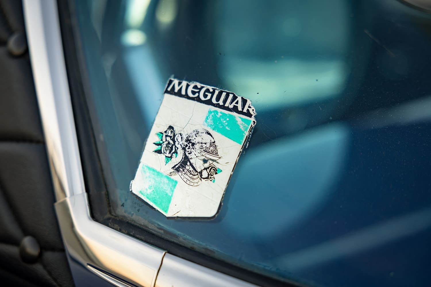 Close-up detail of a weathered vintage Meguiar's helmet and crest decal on the corner of the hardtop window glass.