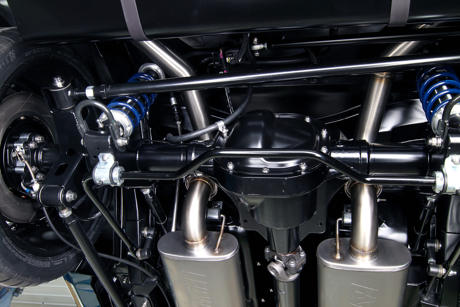 Straight-on underside view of the rear axle assembly, sway bar, rear coilovers, fuel tank, and polished stainless exhaust pipes.