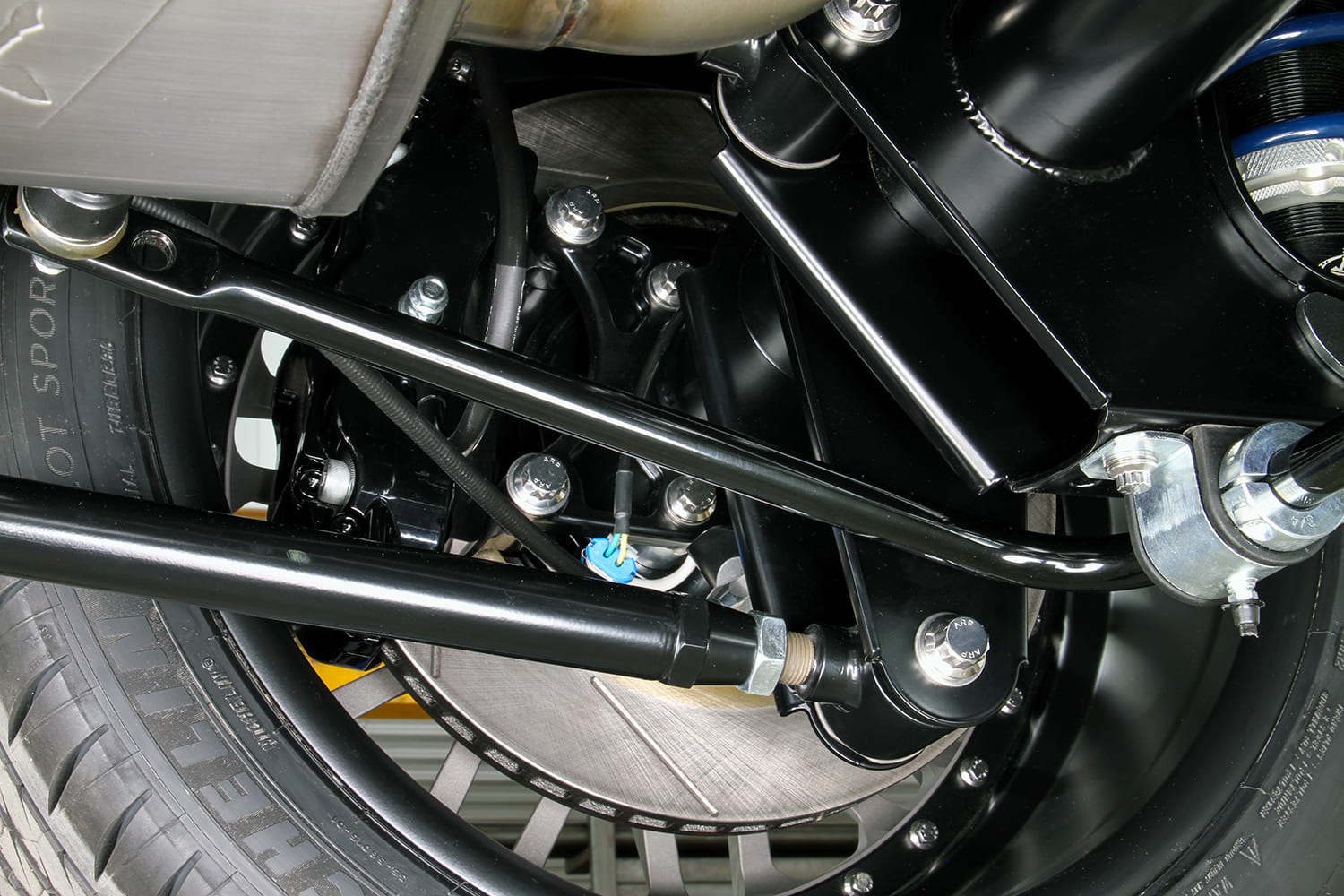 Close-up of the rear suspension trailing arm with heim joint, slotted disc brake rotor, and wide Michelin tire.