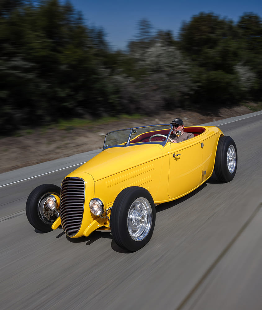 Elevated dynamic highway action shot of a yellow 1934 Ford roadster driving open-air at speed.
