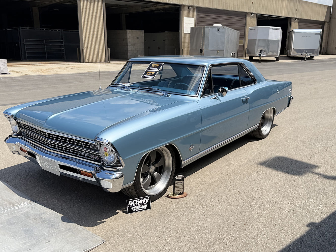 Front three-quarter angle of a light metallic blue 1966 Chevrolet Nova SS sitting low on gunmetal wheels next to its Goodguys award plaque.