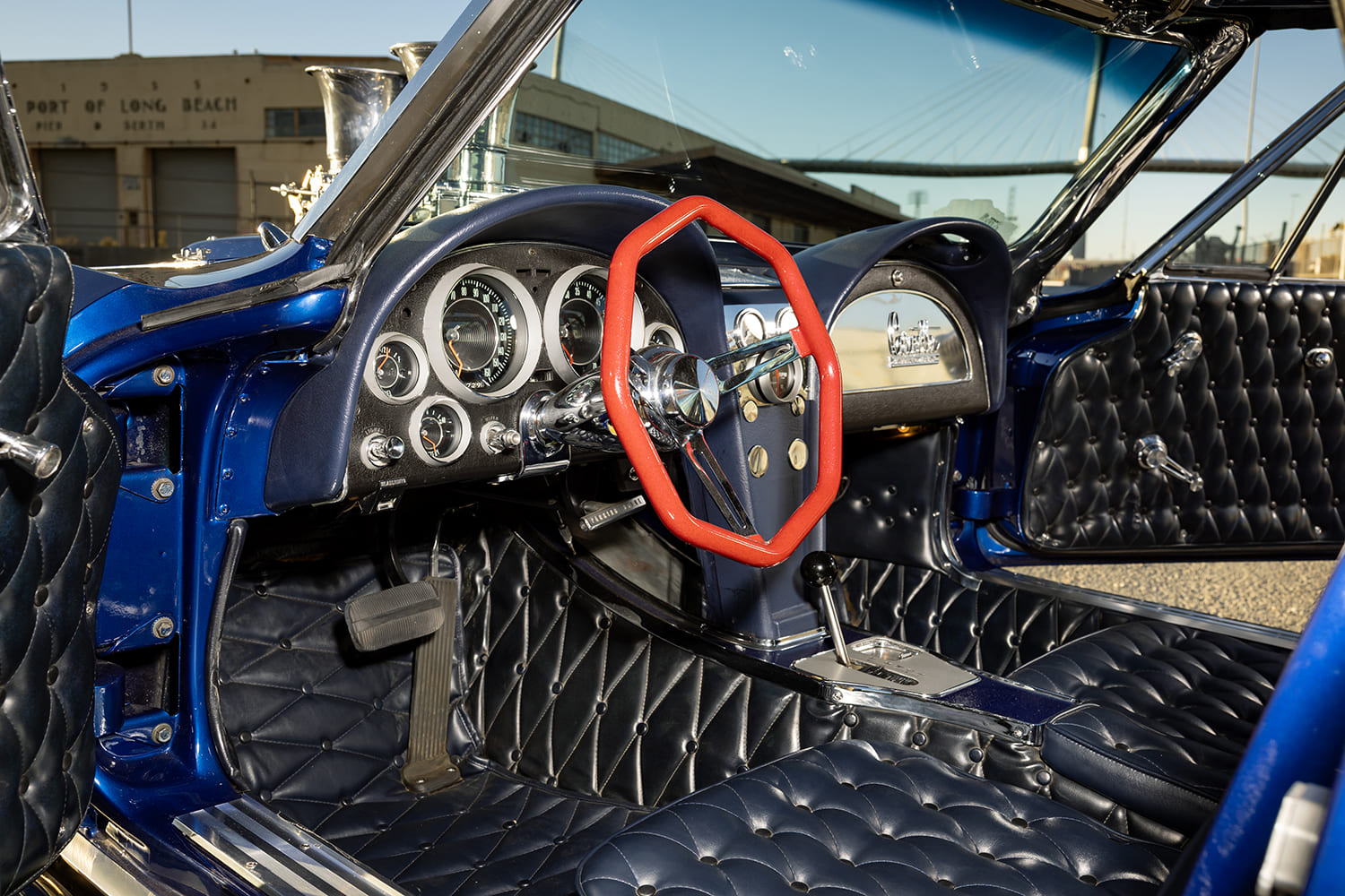 Driver's side view inside the cockpit displaying the red hexagonal steering wheel, factory dash gauges, and deep button-tufted dark blue upholstery.
