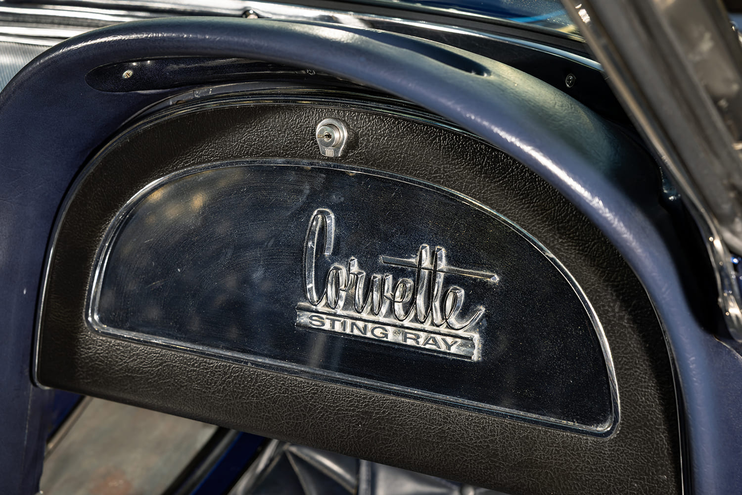 Close-up of the passenger-side dash cove with the polished Corvette Sting Ray emblem and keyed lock on the glovebox door.