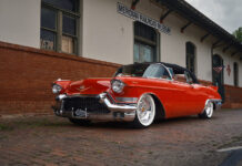 Low front three-quarter view showing the massive chrome front bumper, grille guards, Mike Curtis custom wheels, and Mike Goldman Customs license plate frame.