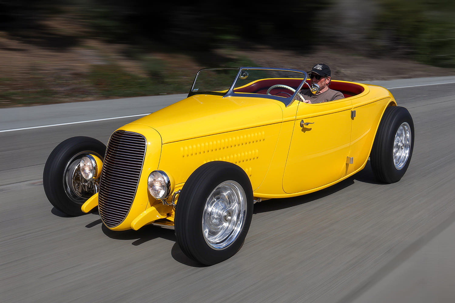 Low tracking action photo of the driver piloting the yellow Brizio-built 1934 Ford roadster on the open road.