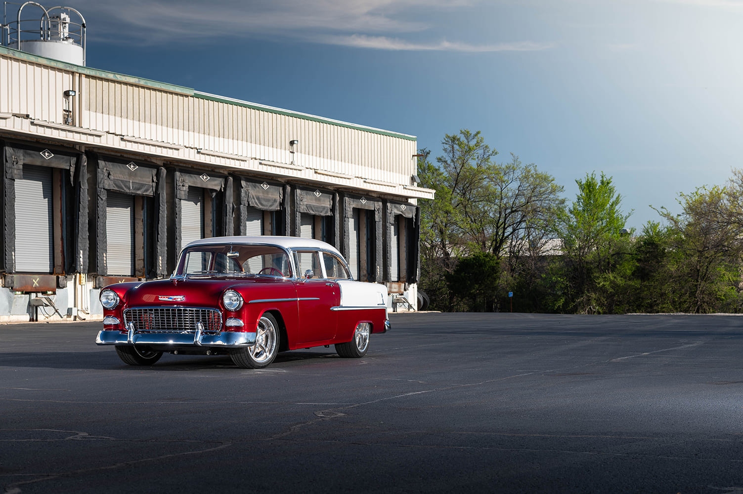 Wide outdoor shot showing the 1955 Bel Air restomod framed against roll-up warehouse bay doors under a sunny sky.