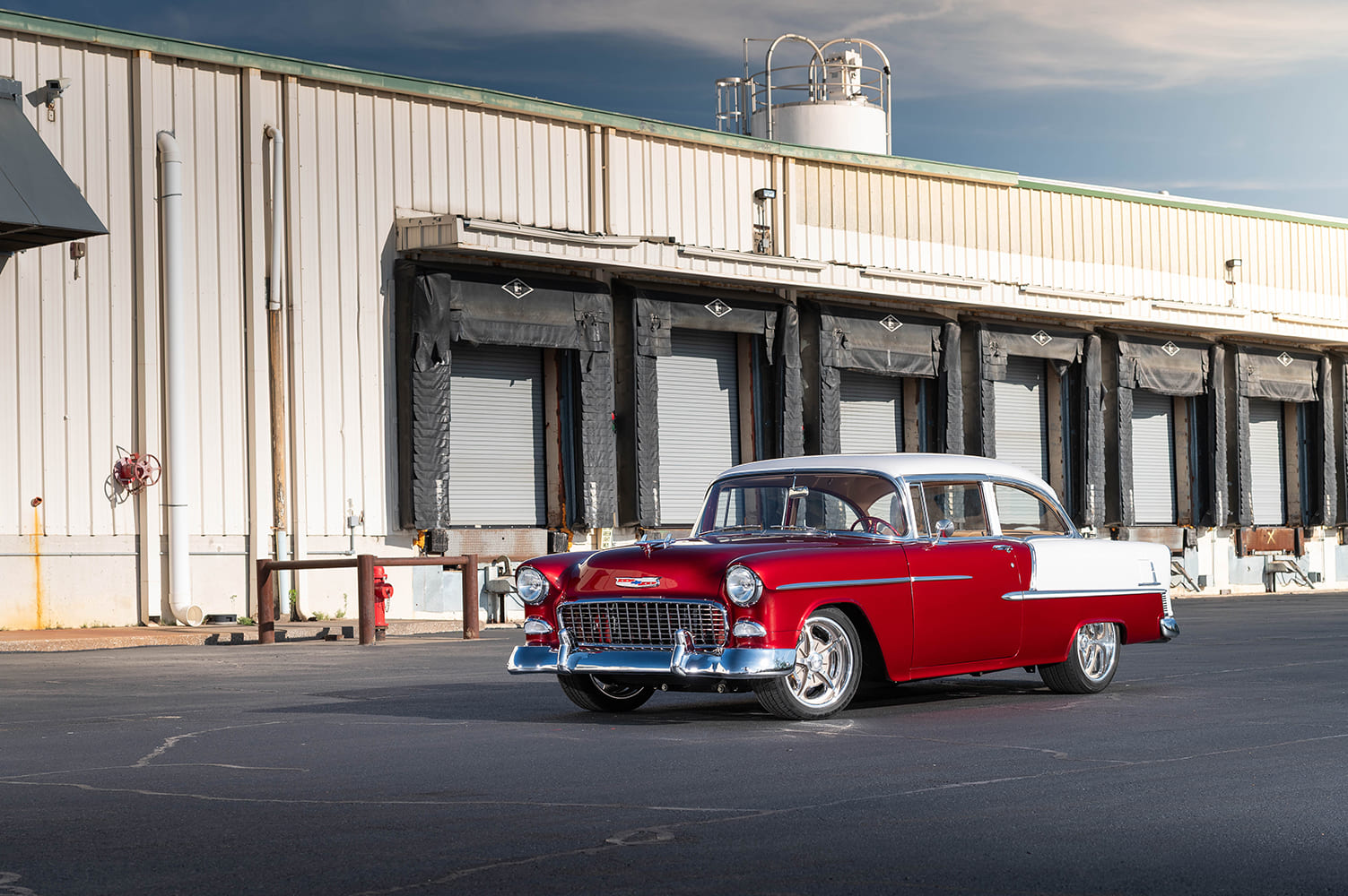 Long-distance front three-quarter view of the candy red and white 1955 Chevrolet sedan positioned in front of an industrial facility and loading bays.