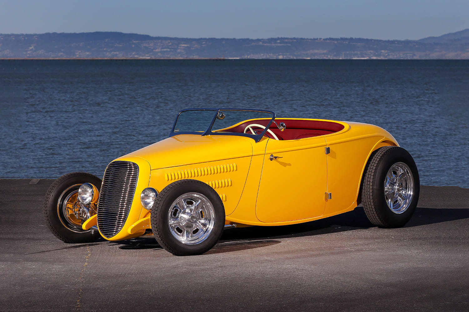 Static three-quarter front view of the yellow Myers 1934 Ford roadster parked along the waterfront bay.