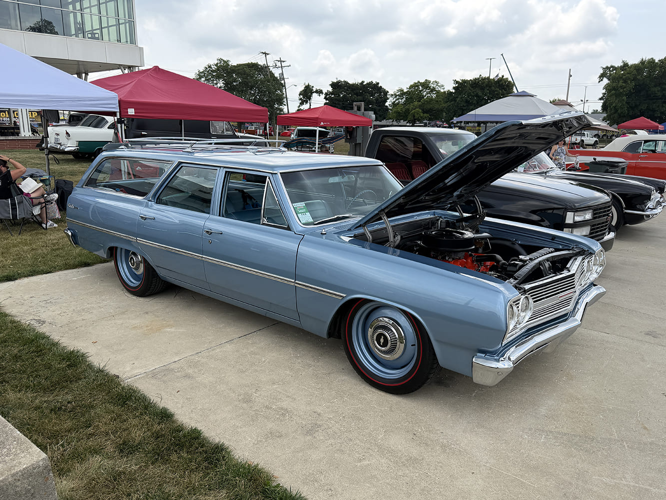 Light blue 1964 Chevrolet Chevelle four-door station wagon with open hood, color-matched steel wheels, and redline tires parked near event tents.