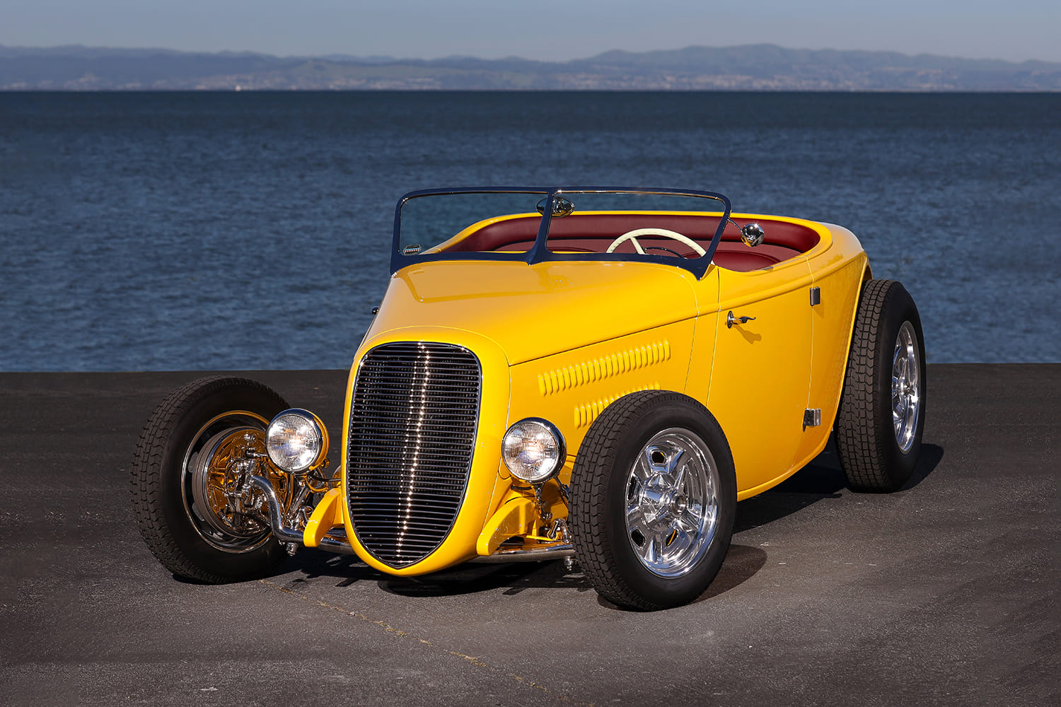 Head-on view showcasing the custom track-style grille, low-mounted headlights, and chrome front axle of the yellow roadster.