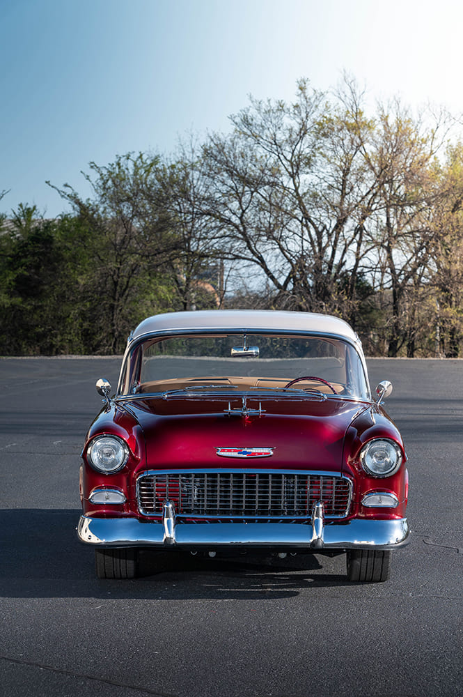 Vertical orientation head-on shot of the 1955 Chevy Bel Air front fascia, chrome bumper, and hood emblem with green trees in the background.