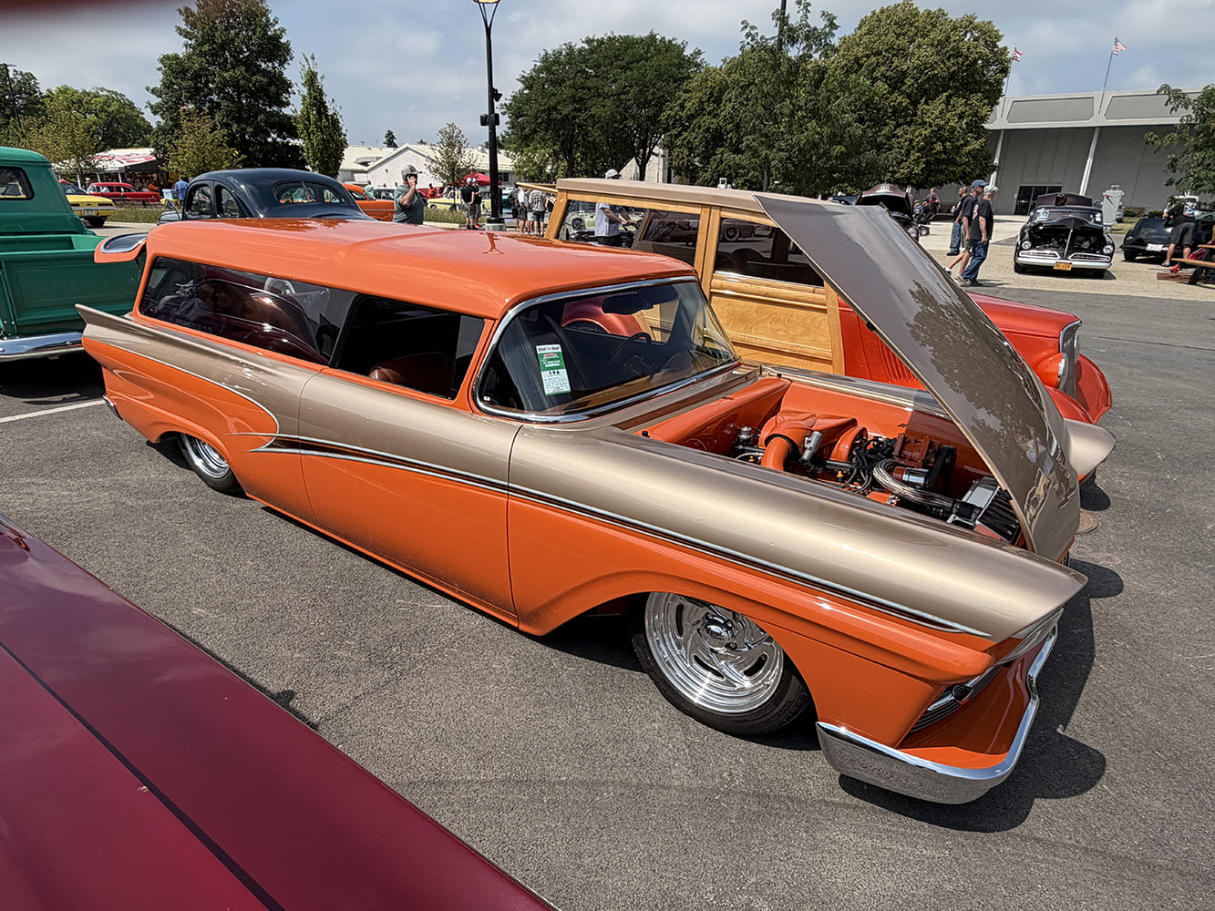 High front three-quarter view of an orange and bronze 1957 Ford custom sedan delivery wagon with tilted open hood in the sunny outdoor lot.