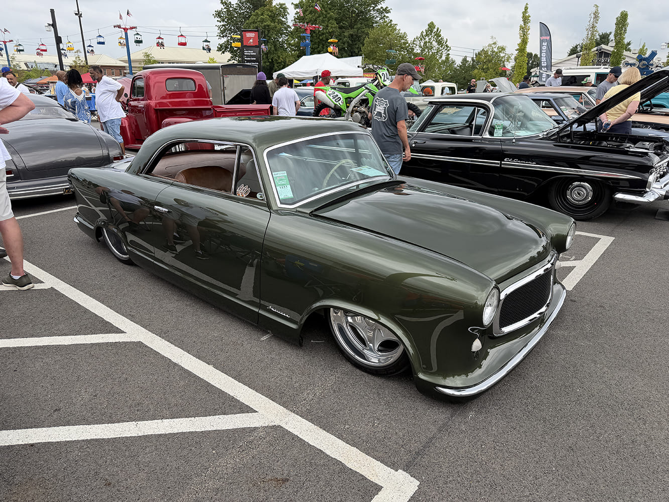 Laid-out dark olive green vintage Rambler American coupe resting low over polished billet wheels in an asphalt parking space.