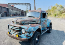 Front three-quarter angle of a weathered blue 1949 Ford F2 4x4 pickup truck with amber fog lights, off-road tires, and "Casey's Rusto-Mods" door lettering parked in an industrial lot.