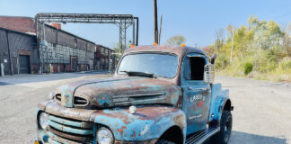 Front three-quarter angle of a weathered blue 1949 Ford F2 4x4 pickup truck with amber fog lights, off-road tires, and "Casey's Rusto-Mods" door lettering parked in an industrial lot.