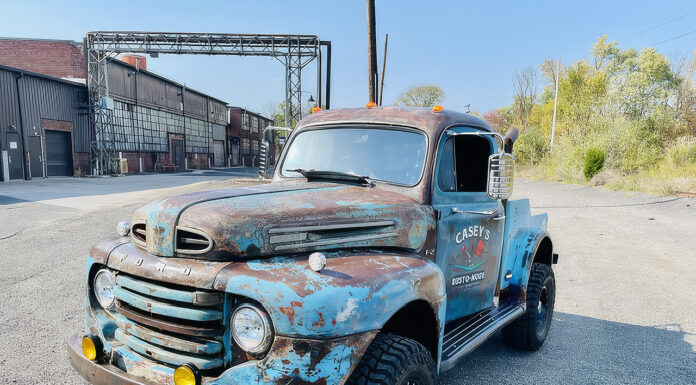 Front three-quarter angle of a weathered blue 1949 Ford F2 4x4 pickup truck with amber fog lights, off-road tires, and "Casey's Rusto-Mods" door lettering parked in an industrial lot.