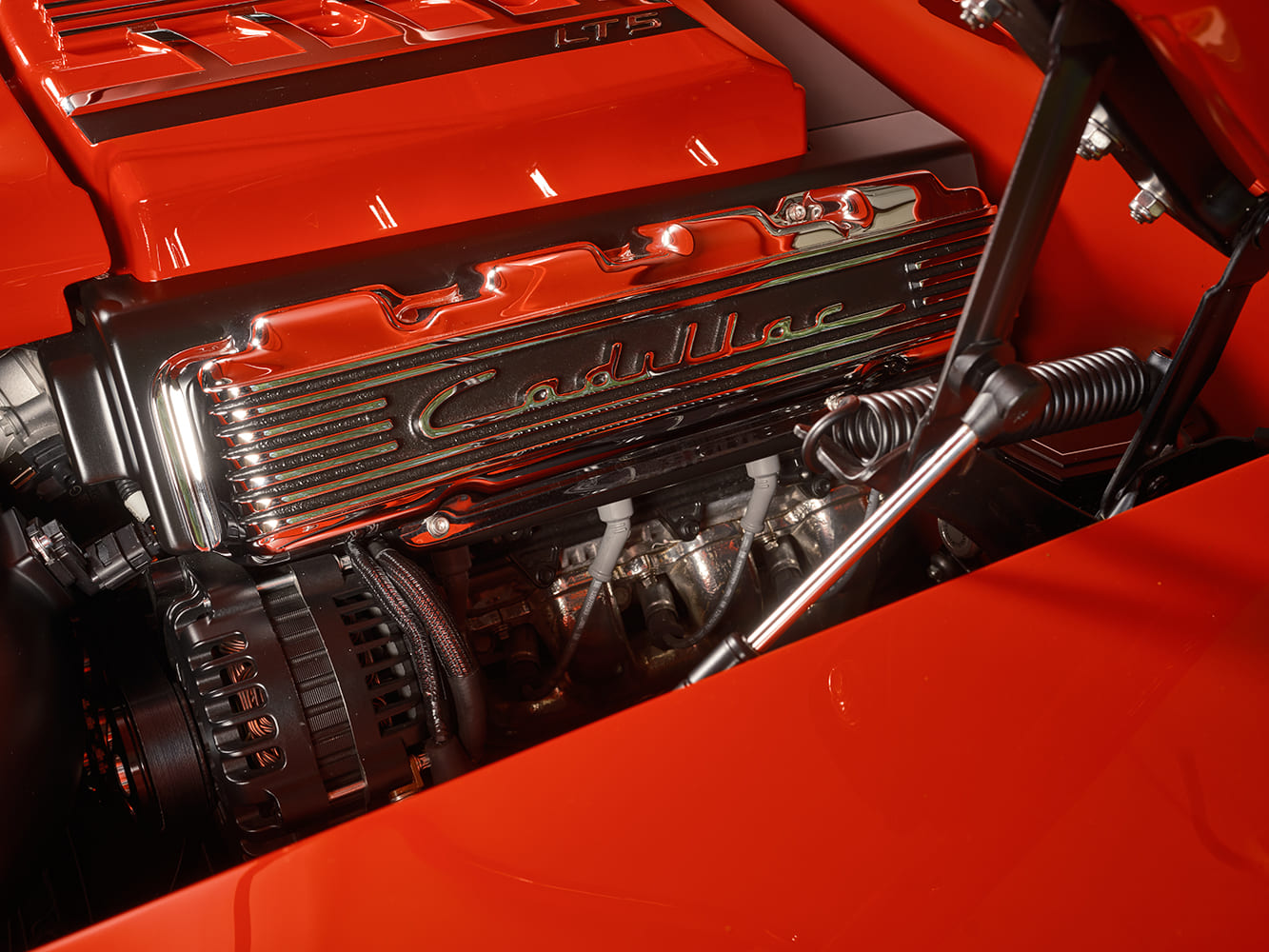 Close-up of the adapted finned chrome Cadillac script valve cover, Wegner serpentine drive pulleys, and Wilwood master cylinder against the slick red firewall.