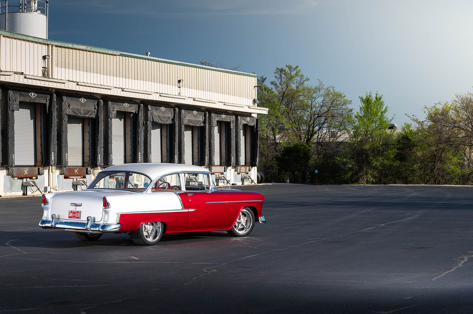 Wide outdoor perspective of the two-tone red and white 1955 Chevrolet Bel Air parked across an empty asphalt lot outside an industrial warehouse.