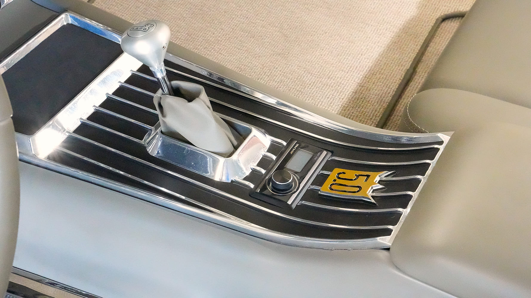 Close-up of the leather-wrapped center console featuring a ribbed aluminum insert, brushed Lokar automatic shifter, flush Alpine controller, and custom Galaxie 5.0 crest.