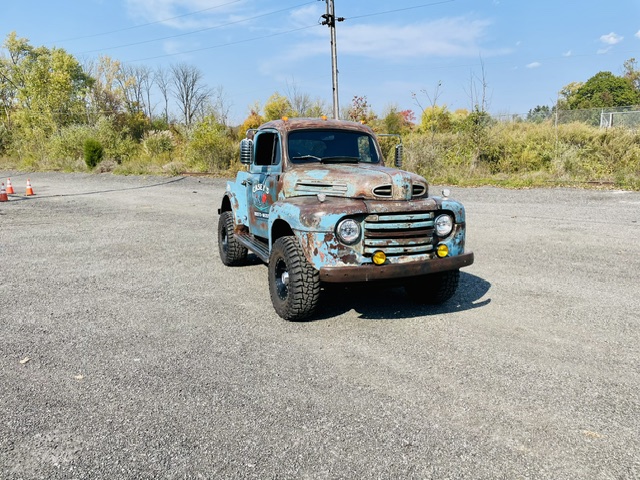 Straight-on front angle of a lifted patina turquoise 1949 Ford F2 pickup truck featuring amber driving lamps, black wheels, and cab clearance lights on gravel.