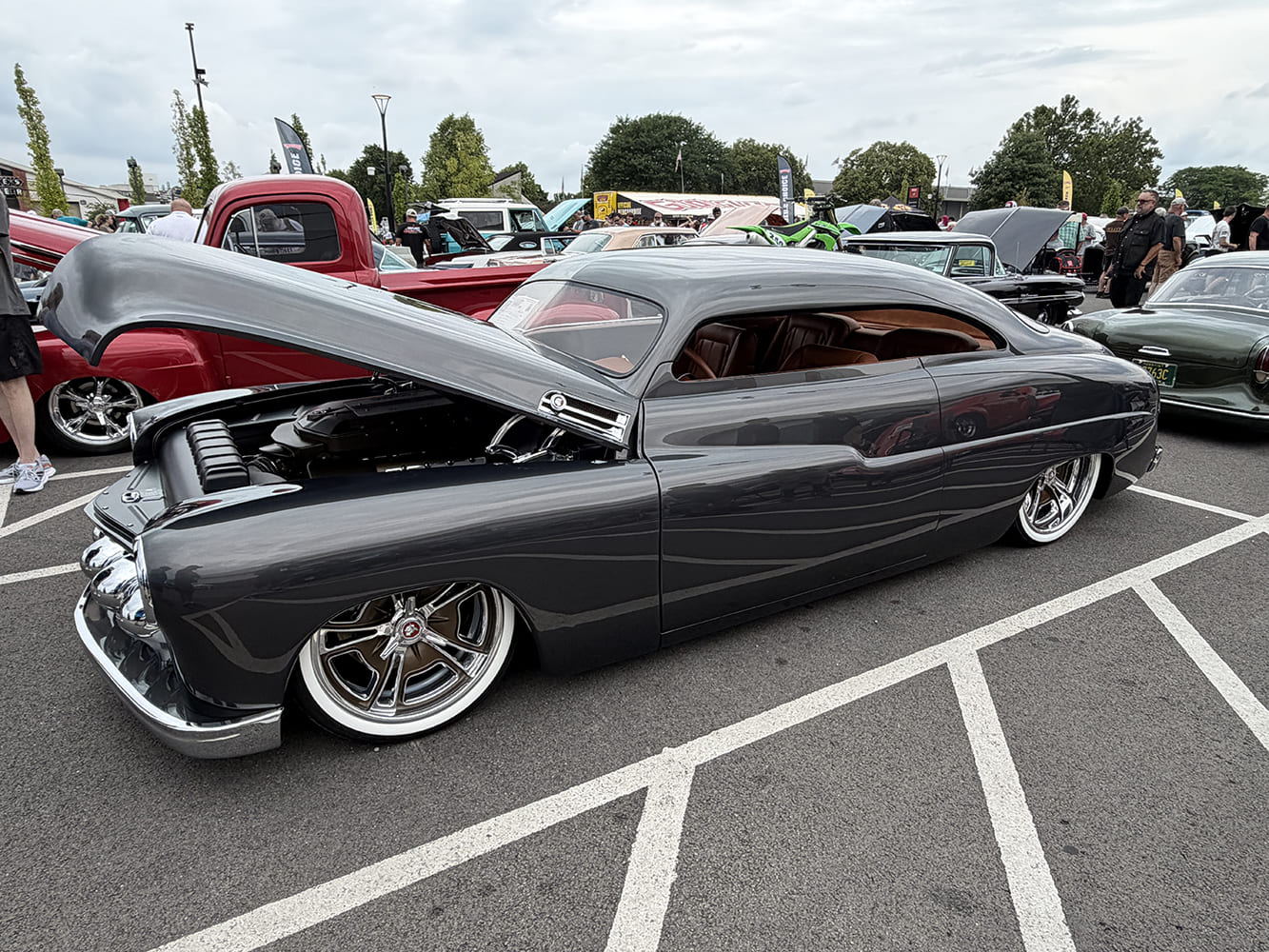 Low-angle side view of a radically chopped and bagged dark gray metallic 1950 Mercury custom coupe with wide whitewall tires and open hood.