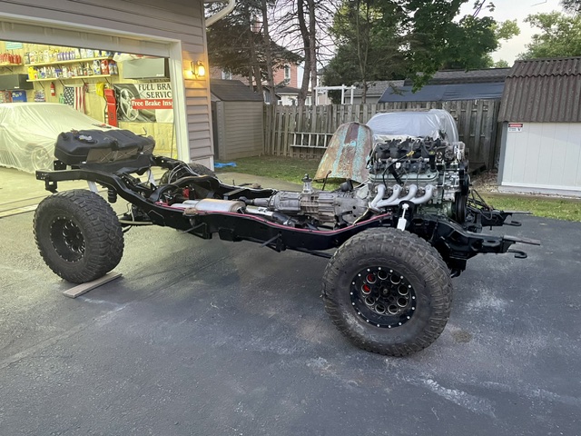 Full side view of the bare 4x4 rolling truck frame equipped with a modern V8 engine, manual transmission, transfer case, and large off-road tires parked in a driveway.
