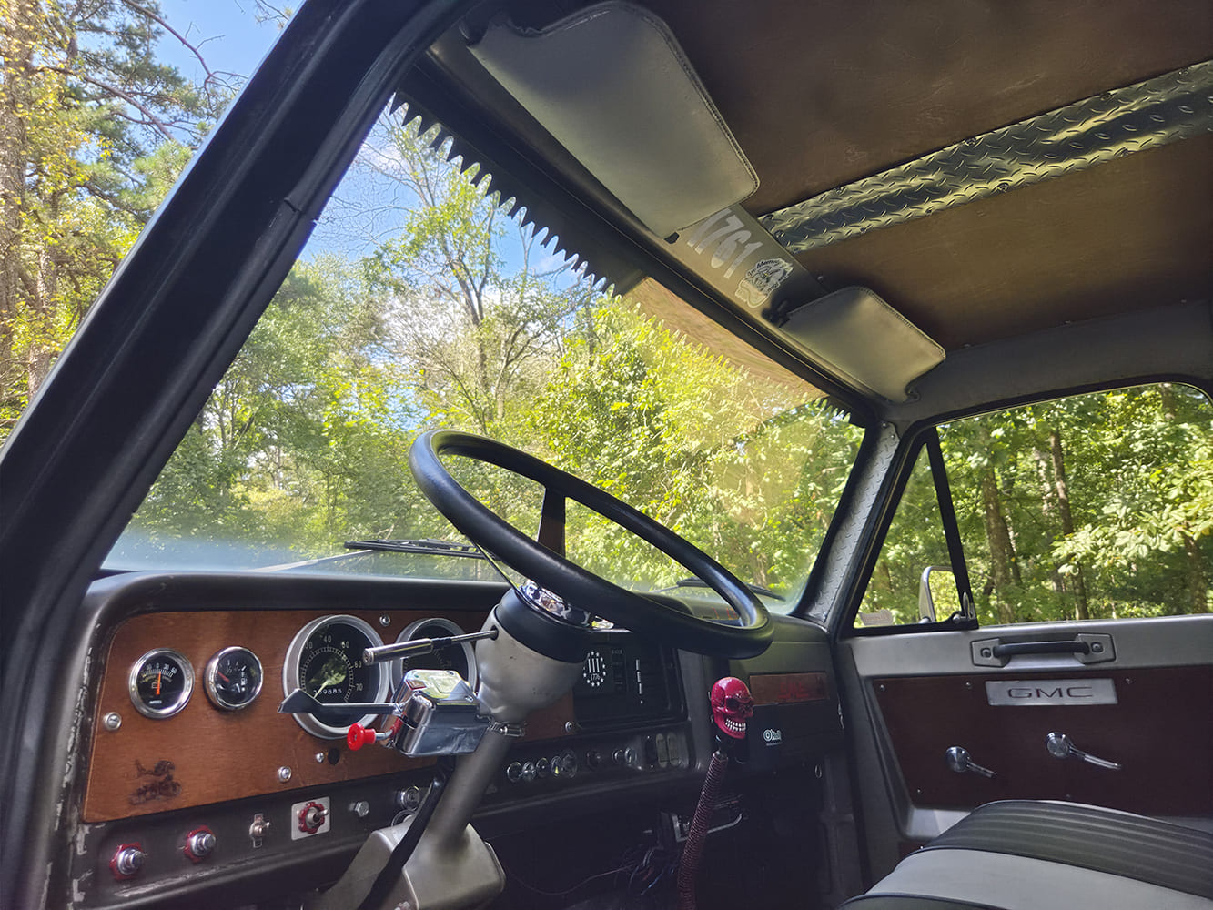 Wide-angle view from the driver side inside a 1967 GMC 7500 showing wood-grain dash face, round gauges, and custom overhead headliner.