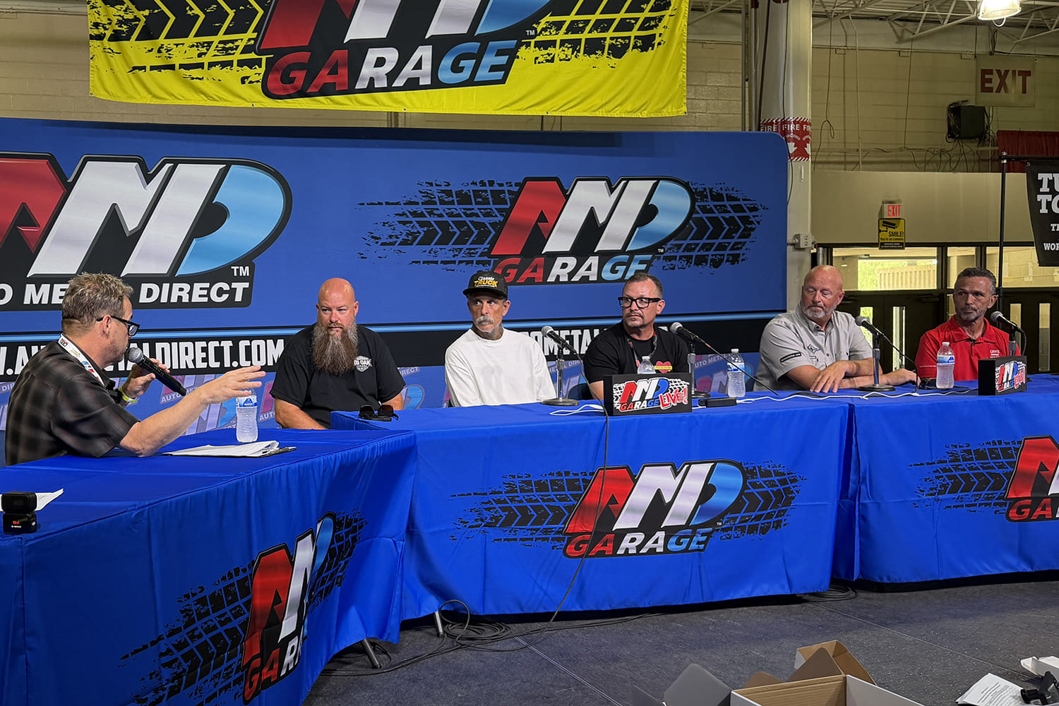 Automotive builders and industry figures seated at the Auto Metal Direct AMD Garage Live panel discussion table speaking into microphones.