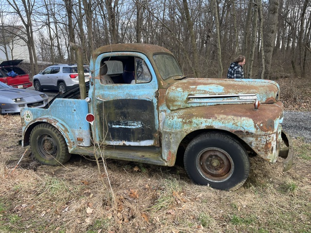 Driver-side profile of the weathered blue 1949 Ford F2 truck sitting derelict in a wooded field prior to restoration, showing heavy rust and a vintage vertical exhaust stack.
