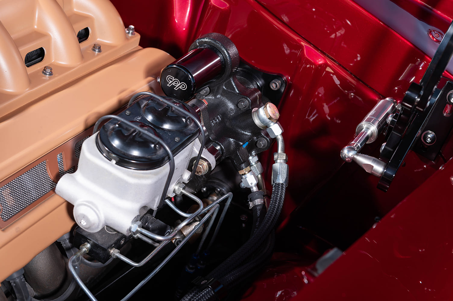 Detailed close-up of a black CPP hydroboost brake assist unit with hardlines mounted to the deep metallic red firewall next to billet hood hinges.