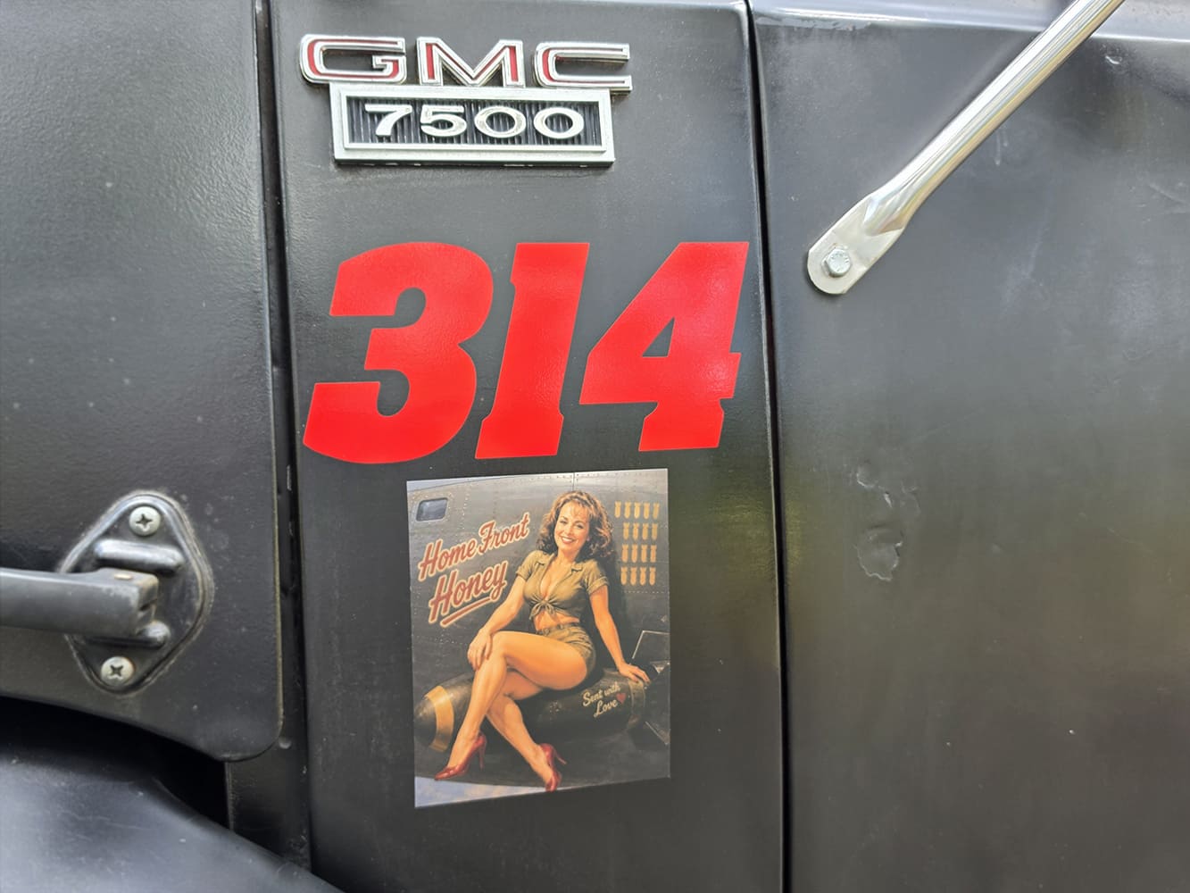 Close-up of original chrome GMC 7500 fender emblem above red 314 numbers and a vintage WWII-style "Home Front Honey" pin-up decal.