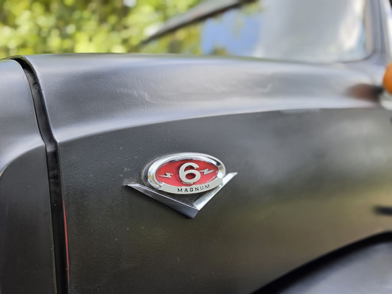 Close-up of red and chrome "V-6 Magnum" hood side badge mounted on the matte black bodywork of a 1967 GMC 7500.