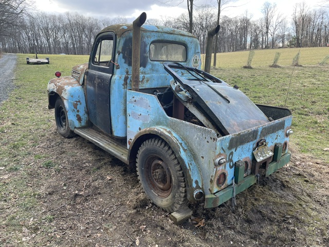 Rear three-quarter view of the unrestored 1949 Ford F2 wrecker resting on overgrown grass with weathered paint, dual exhaust stacks, and a manual hand-crank boom frame.