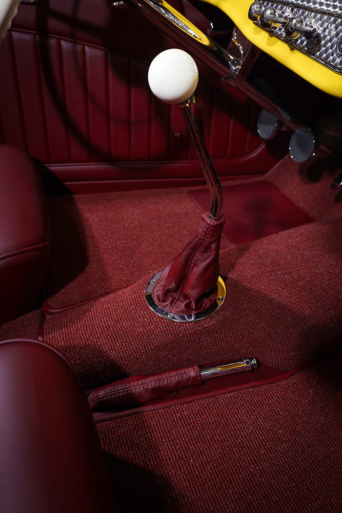 Close-up of the curved chrome floor shifter, red leather shift boot, white shift ball, and matching emergency brake lever on red loop carpet.