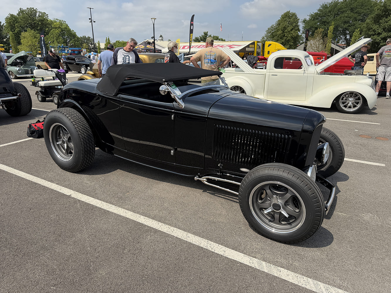 Front three-quarter view of a clean gloss black 1932 Ford highboy roadster with black soft top, open hood sides, and five-spoke gray wheels on outdoor asphalt.
