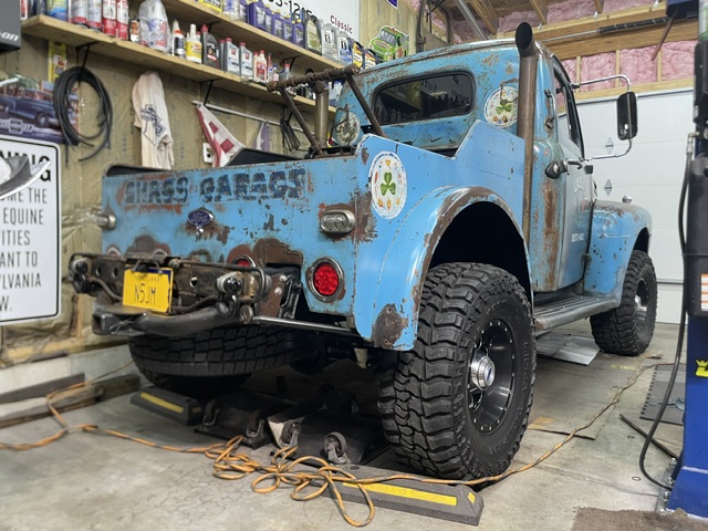 Low rear three-quarter shop view of the lifted 1949 Ford F2 showing the under-bed spare tire mount, heavy-duty tow shackle bracket, and aggressive mud-terrain tires.