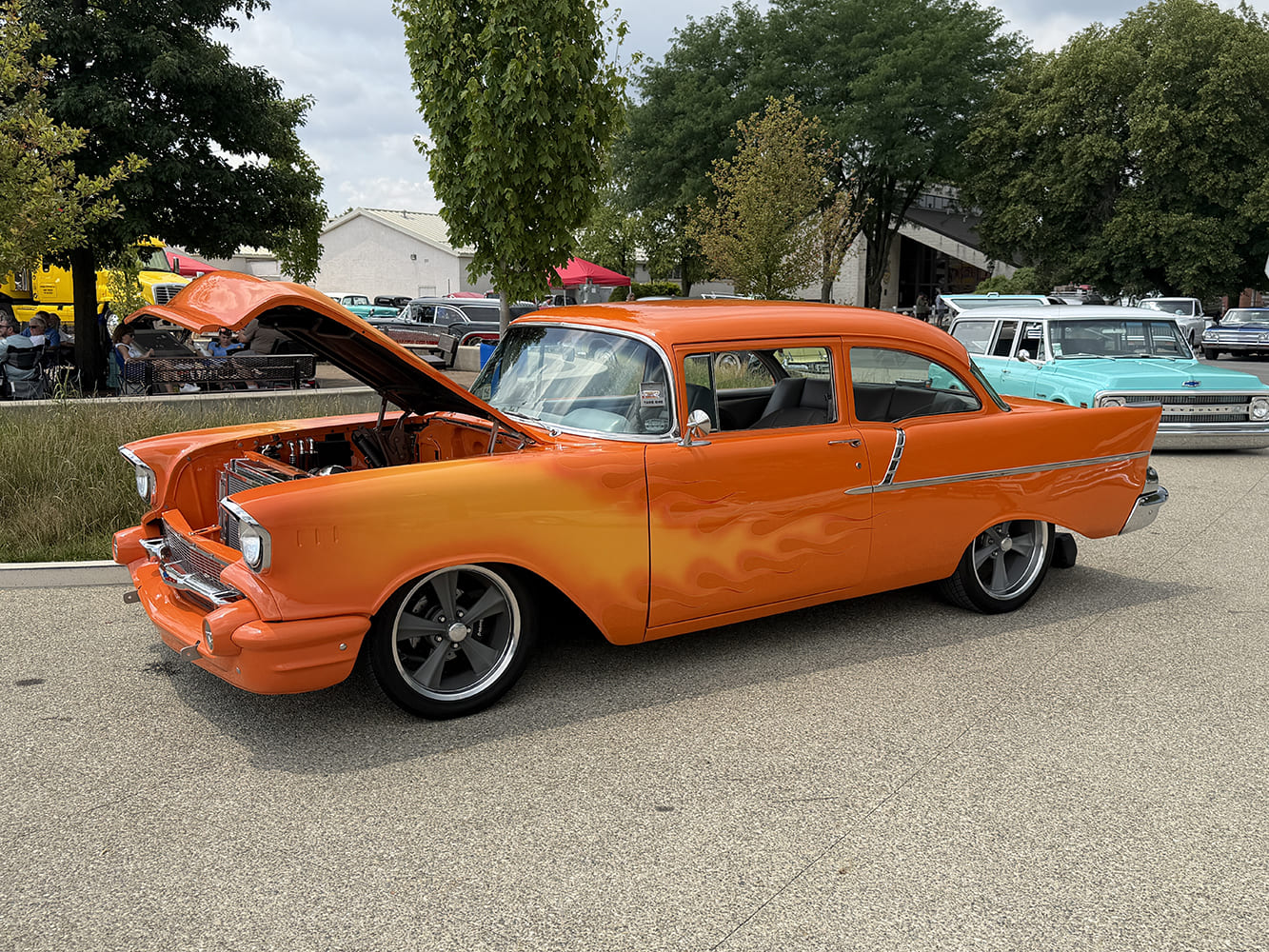 Side view of a lowered bright orange 1957 Chevrolet two-door post sedan featuring subtle yellow ghost flames and gray American Racing Torq Thrust wheels.