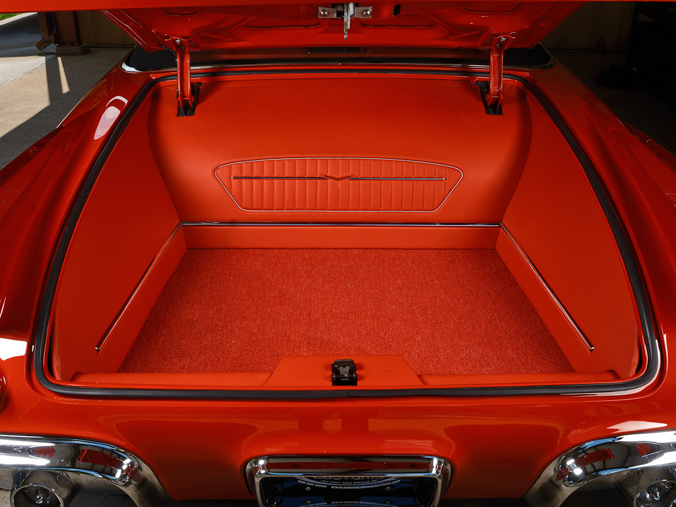 Wide-angle view into the open trunk featuring smooth body-color panels, pleated leather insert with Cadillac emblem, and matching red carpet.
