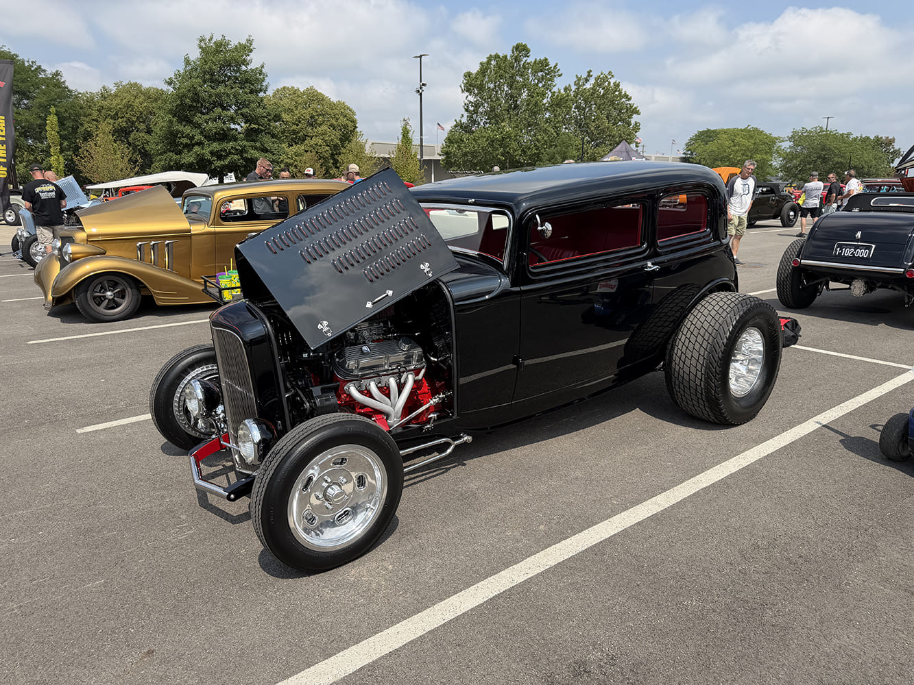 Front three-quarter view of a channeled gloss black 1932 Ford Tudor sedan with an open louvered hood top, red engine block, and wide rear tires.