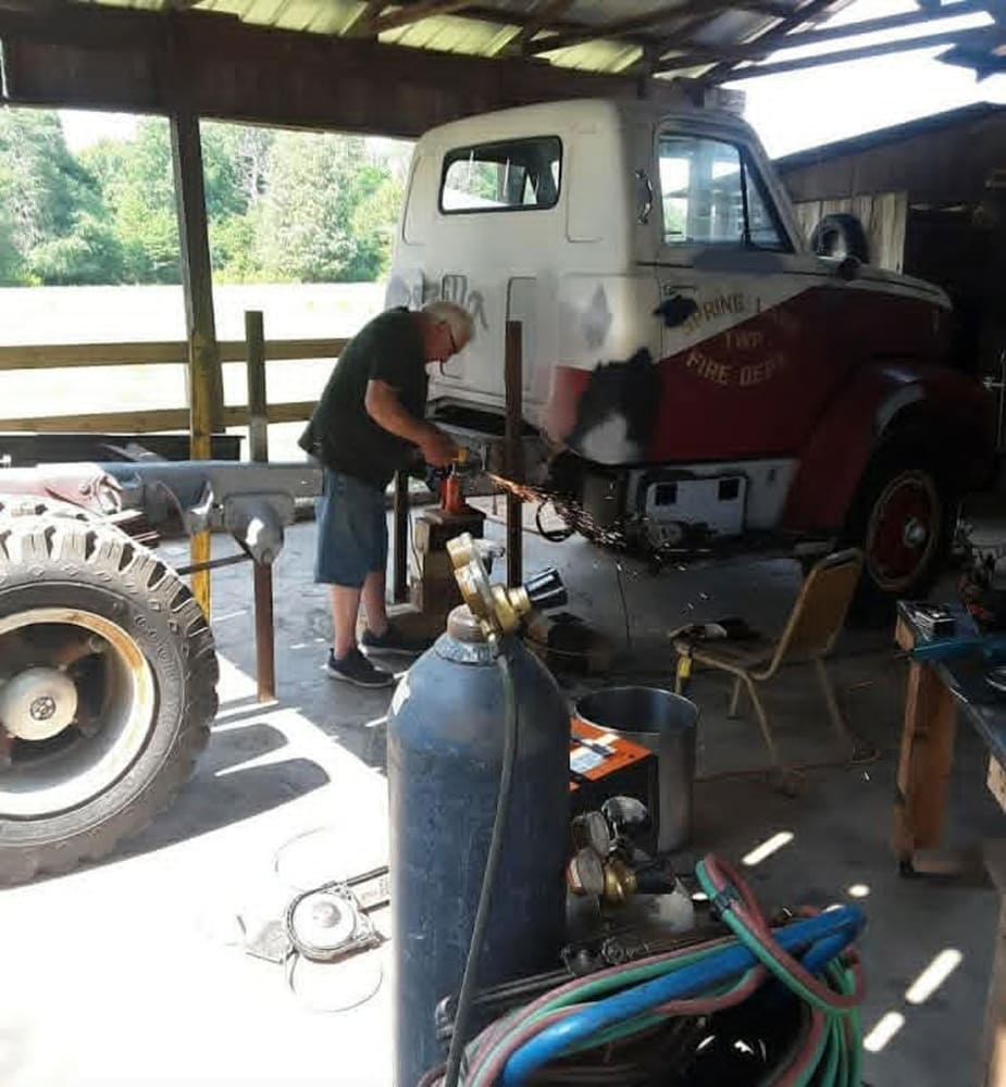 Workshop view of builder grinding and fabricating the cab and chassis of a vintage GMC 7500 medium-duty fire truck donor.