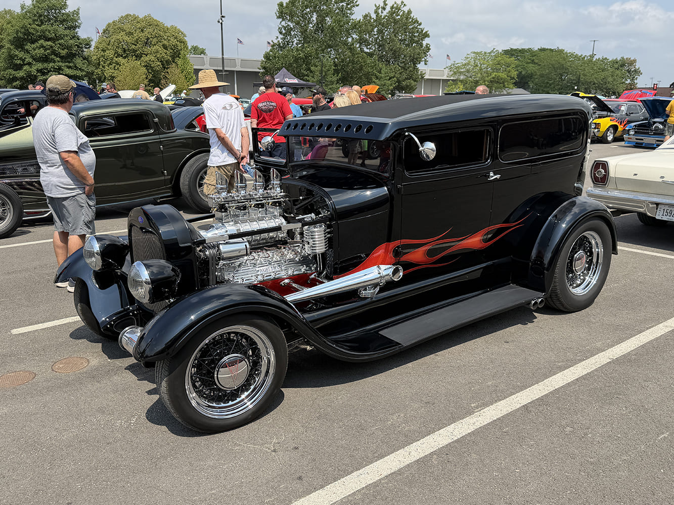 Front three-quarter view of a black 1929 Ford Model A Tudor sedan hot rod featuring orange flames, an exposed supercharged V8, and wire wheels parked in an event lot.